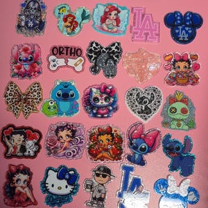Puede incluir: Una colección de dijes decorativos con detalles brillantes. Los diseños incluyen personajes de dibujos animados como Stitch, Betty Boop y Hello Kitty, junto con logotipos y lazos. Los dijes son de varios colores, incluyendo rosa, azul y negro.
