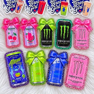 May include: A collection of twelve rectangular and heart-shaped stickers featuring various energy drink cans and Dutch Bros branding. The stickers are decorated with glittery borders and satin bows in pink, green, and blue, showcasing colorful designs.