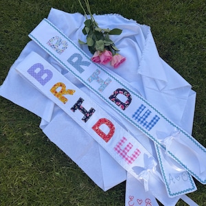 May include: Three white sashes with the word "BRIDE" spelled out in colorful, patterned fabric letters. The sashes are decorated with a light blue zigzag trim. Two small white ribbons are also visible, embroidered with initials.
