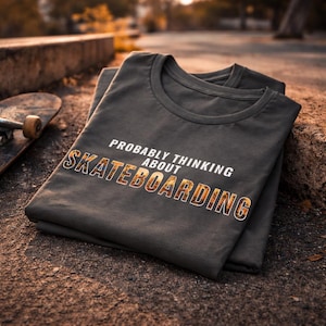 Könnte beinhalten: Zwei dunkelgraue T-Shirts sind gefaltet und gestapelt, wobei das obere Shirt den Text "PROBABLY THINKING ABOUT SKATEBOARDING" in weißen und orangefarbenen Buchstaben trägt. Im Hintergrund ist ein Skateboard zu sehen, was auf ein Skateboarding-Thema hindeutet.