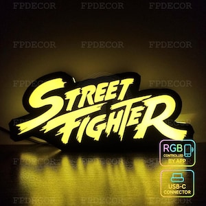 Op de afbeelding: Een neonbord met de tekst "Street Fighter" in heldergele letters, omlijnd met zwart. Het bord is verlicht en bevat pictogrammen die RGB-bediening via een app en een USB-C-connector aangeven.