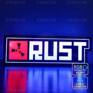 Pode incluir: Uma placa LED retangular com a palavra "RUST" em letras brancas iluminadas. O lado esquerdo apresenta um quadrado vermelho com um gráfico preto. A placa inclui ícones "RGB controlado por aplicativo" e "Conector USB-C".