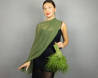 Può includere: Una donna indossa un abito nero e uno scialle verde oliva. Tiene in mano una borsa abbinata ornata di piume verdi. Lo sfondo è grigio.