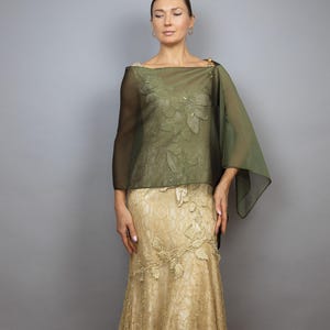 Puede incluir: Una mujer lleva un vestido de encaje dorado con una estola de gasa verde oliva. La estola tiene un diseño floral y está drapeada sobre sus hombros.