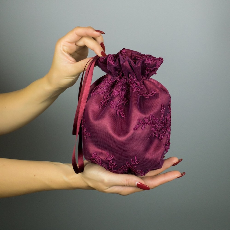 May include: A deep burgundy satin drawstring bag with floral lace embroidery. The bag has a gathered top and is held by hands, highlighting the colour and texture. The drawstring ribbons are a matching burgundy.