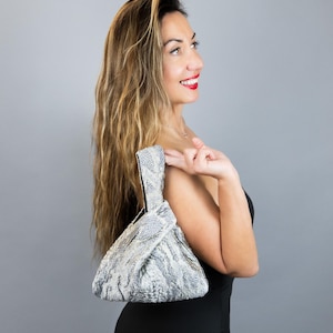 Puede incluir: Un pequeño bolso triangular con un estampado de piel de serpiente en tonos grises y blancos. El bolso tiene un asa anudada y un interior negro. La mujer de la imagen lleva un vestido negro.