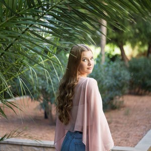 May include: A woman with long, wavy brown hair is wearing a light pink sheer top and blue jeans. She is standing in front of a palm tree and looking over her shoulder.