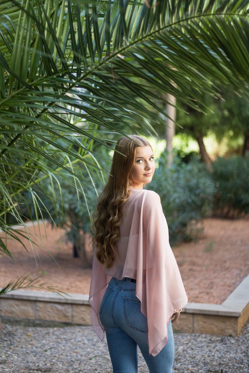 May include: A woman with long, wavy brown hair is wearing a light pink sheer top and blue jeans. She is standing in front of a palm tree and looking over her shoulder.