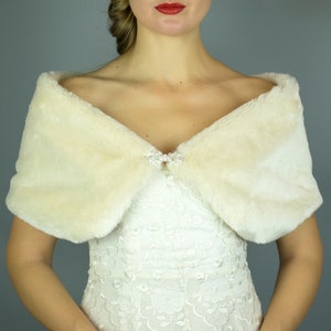 May include: A white faux fur shawl with a crystal brooch. The shawl is draped over the shoulders of a person wearing a white lace dress.