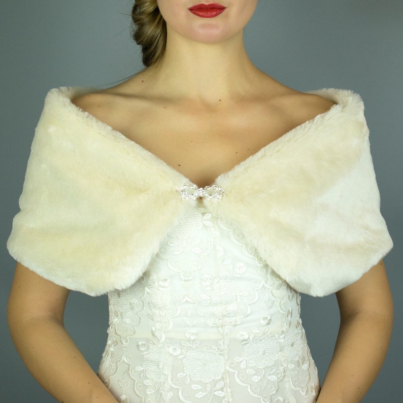 May include: A white faux fur shawl with a crystal brooch. The shawl is draped over the shoulders of a person wearing a white lace dress.