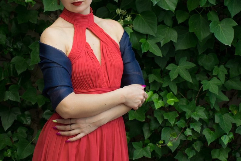 May include: A woman wearing a red halter dress with a navy blue shawl draped over her shoulders. She is standing in front of a green leafy wall.