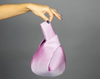 May include: A light purple satin handbag with a knotted handle. The bag has a rounded, teardrop shape and is held up by a hand against a gray background. The satin fabric has a subtle sheen.