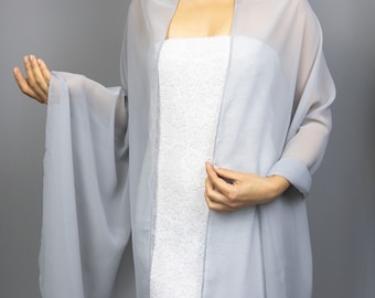 Puede incluir: Un chal transparente gris claro sobre un vestido blanco. El chal tiene mangas largas y un diseño fluido. El vestido parece estar adornado con lentejuelas o cuentas blancas. El fondo es gris liso.