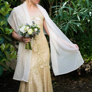 May include: A bride wearing a gold lace wedding dress with a white chiffon shawl. She is holding a bouquet of white and green flowers.
