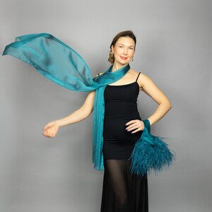 May include: A teal scarf and a matching wrist accessory with feathers. The scarf is draped around the neck and flowing. The woman is wearing a black dress.