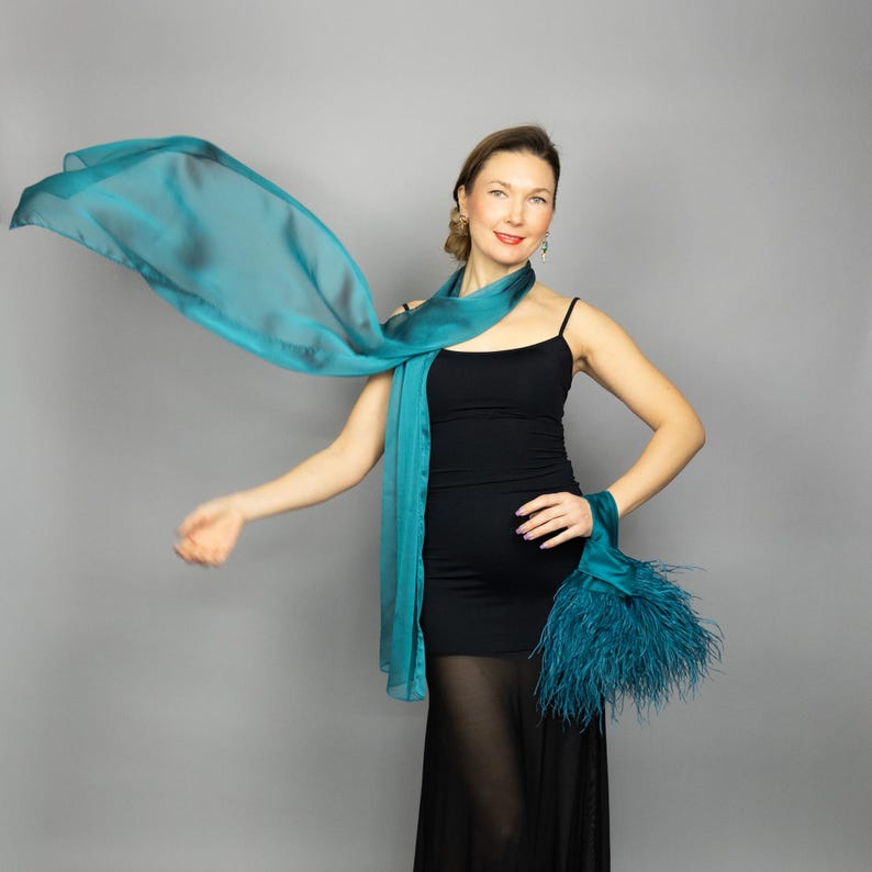 May include: A teal scarf and a matching wrist accessory with feathers. The scarf is draped around the neck and flowing. The woman is wearing a black dress.