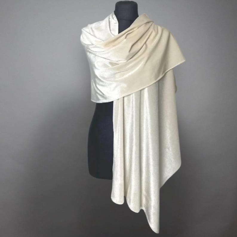 May include: A cream-colored velvet shawl draped over a black mannequin. The shawl is soft and flowing, with a draped design. The fabric has a subtle sheen, and the overall look is elegant and sophisticated. The shawl is a versatile accessory.