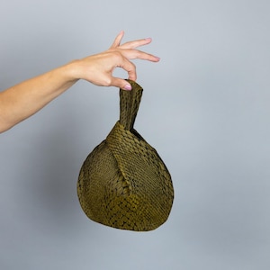 May include: An olive green clutch bag with a textured, scale-like pattern. The bag has a knotted handle and a rounded, triangular shape. The clutch is held up against a grey background.
