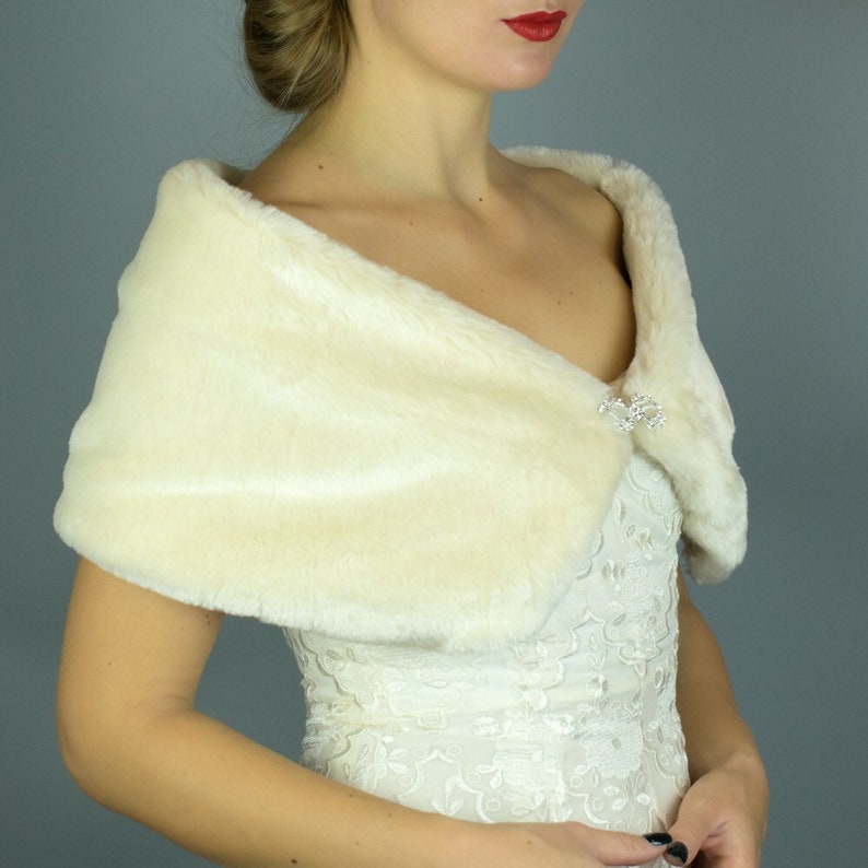 May include: A white faux fur shawl with a crystal brooch. The shawl is draped over the shoulders of a person wearing a white lace dress.