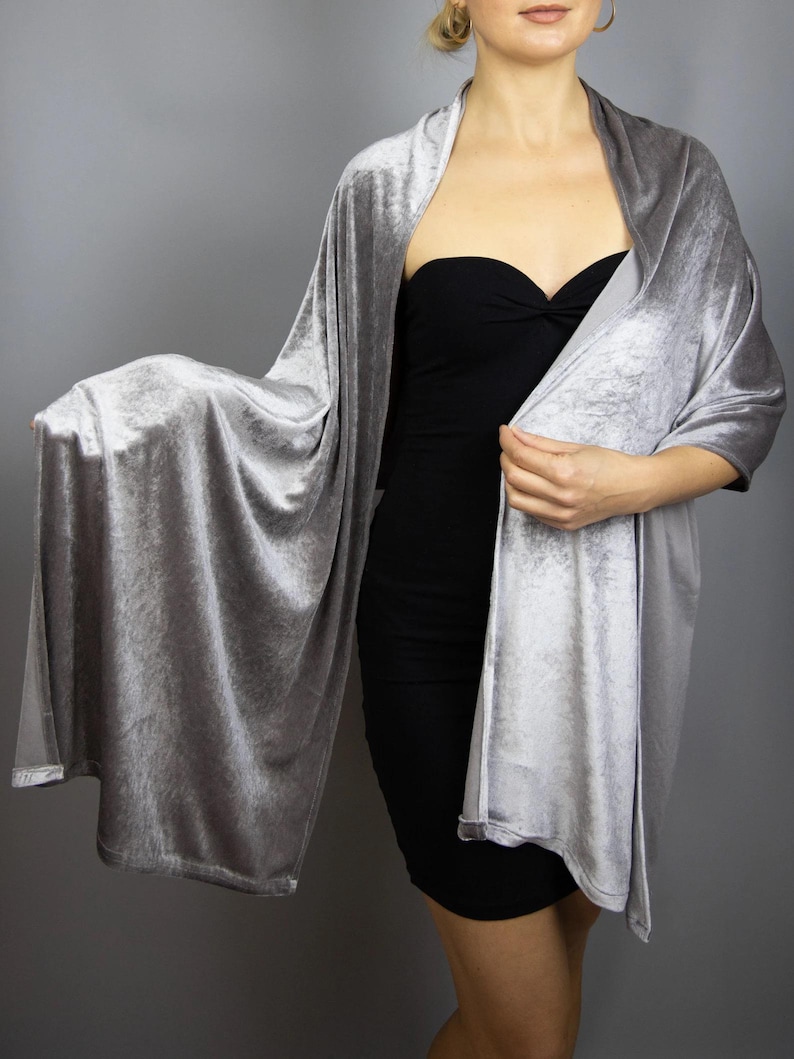 May include: A silver-gray velvet shawl draped over a black dress. The shawl has a soft, flowing texture and drapes over the shoulders and arms. The fabric has a subtle sheen, reflecting light. The overall look is elegant and sophisticated.