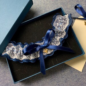 Peut inclure: Une jarretière en dentelle bleue et blanche avec un nœud en satin bleu et un charme d'abeille dorée. La jarretière est dans une boîte bleue avec une étiquette blanche.