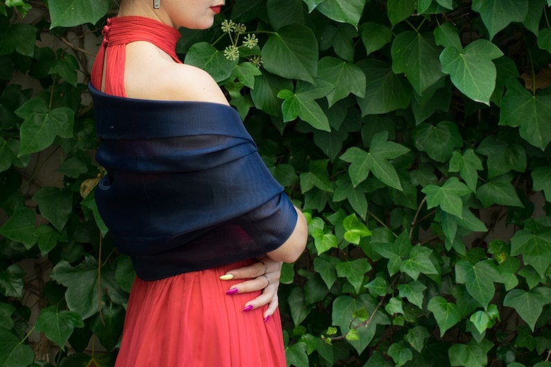 May include: A woman wearing a red dress with a navy blue shawl draped over her shoulders. She is standing in front of a wall covered in green ivy.