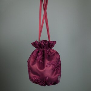 May include: A deep burgundy satin drawstring bag with embroidered floral lace. The bag has a gathered top and is suspended by two matching burgundy satin ribbons. The bag is a rich shade of burgundy.
