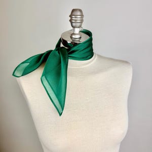May include: A vibrant emerald green scarf is draped around a mannequin's neck. The scarf is made of a sheer fabric, with a square shape and a knotted design. The scarf's color is a rich, deep green, adding a pop of color to any outfit.