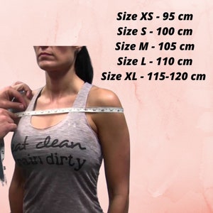 May include: A woman is wearing a grey tank top with the text "eat clean train dirty" on it. She is being measured for size with a tape measure across her shoulders. The text above her reads: "Size XS - 95 cm, Size S - 100 cm, Size M - 105 cm, Size L - 110 cm, Size XL - 115-120 cm."