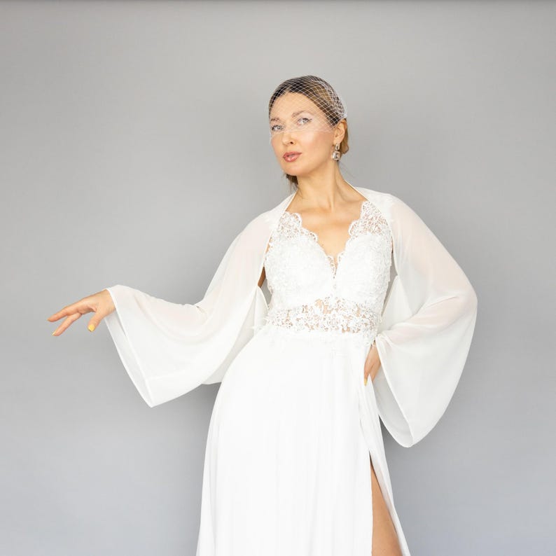 May include: A white bridal gown with a lace bodice and a sheer chiffon overlay. The gown has a long skirt with a slit and a flowing train. The bride is wearing a white veil and has her hair pulled back.