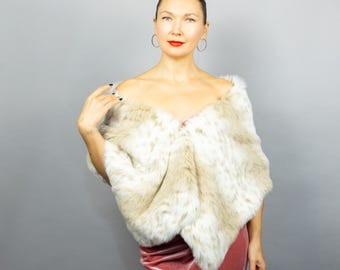 May include: A faux fur shawl in cream and tan tones. The shawl is draped over the shoulders of a person wearing a pink velvet dress. The background is a solid gray.