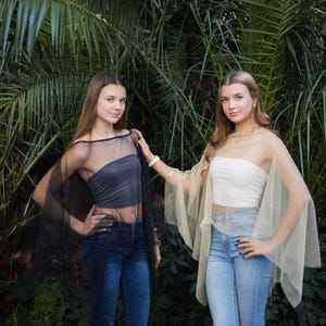 May include: Two women wearing sheer tops and jeans pose in front of lush green foliage. One woman wears a black sheer top over a gray tube top, the other a beige sheer top over a white tube top. Both wear blue jeans.