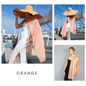 May include: A wide-brimmed orange sun hat and a flowing peach-coloured scarf. The image also features a white dress and the word "ORANGE" in a bold font. The background includes boats and a blue sky.