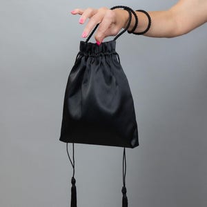 Può includere: Una borsa nera in raso con coulisse e nappe abbinate. È tenuta da una mano con smalto rosa. Il polso ha braccialetti di corda neri. Un accessorio piccolo ed elegante.
