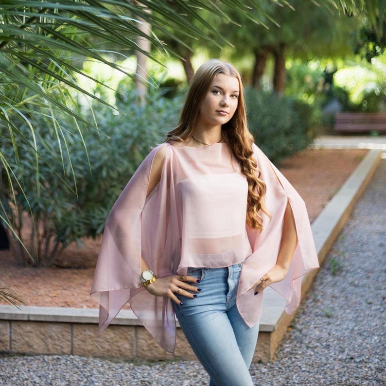 May include: A woman wearing a light pink sheer top with cut-out shoulders and long sleeves. She is wearing blue jeans and has her hands on her hips.