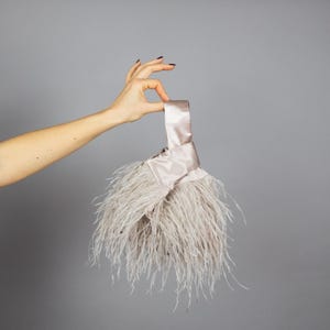 May include: A light grey clutch bag with a satin handle and a fluffy feather design. The bag is held up by a hand against a grey background. The feathers are long and soft, creating a unique texture.