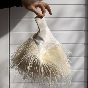 May include: A cream-coloured clutch bag with a handle, decorated with fluffy feathers. The bag has a satin-like texture and is held up by a hand against a white background. The feathers cascade from the bottom of the bag.