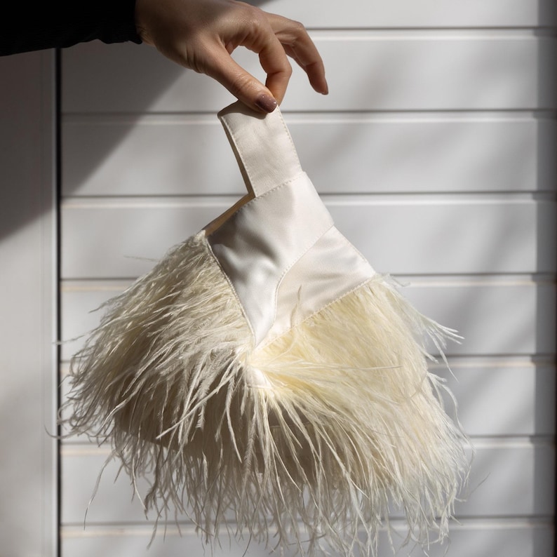 May include: A cream-coloured clutch bag with a handle, decorated with fluffy feathers. The bag has a satin-like texture and is held up by a hand against a white background. The feathers cascade from the bottom of the bag.