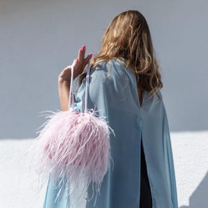 May include: A light pink drawstring pouch bag covered in long, soft feathers. The bag features two thin white braided rope handles. The feathers are pale pink and create a voluminous, textured appearance. The bag is held by a hand.