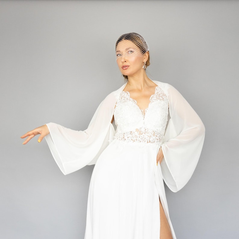 May include: A white bridal gown with a lace bodice and a sheer chiffon overlay. The gown has a long skirt with a slit and a flowing train. The bride is wearing a white veil and has her hair pulled back.