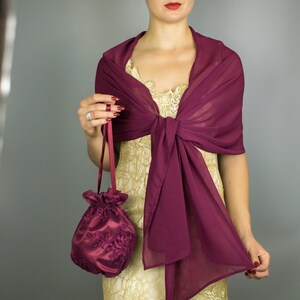 May include: A burgundy chiffon shawl draped over a gold dress. A matching burgundy satin drawstring bag with floral embroidery is held. The shawl is knotted at the front. The overall theme is formal wear.