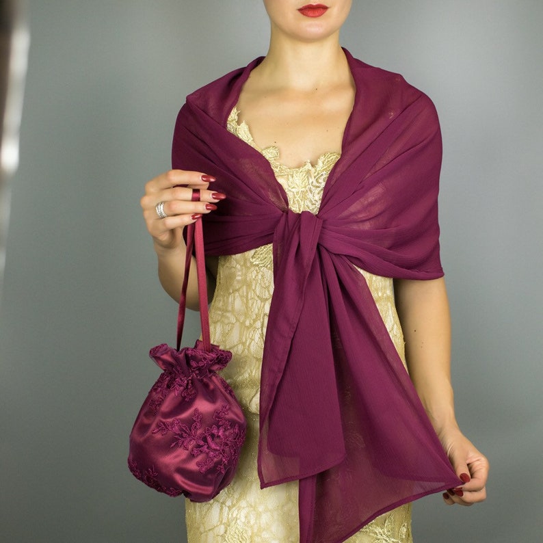May include: A burgundy chiffon shawl draped over a gold dress. A matching burgundy satin drawstring bag with floral embroidery is held. The shawl is knotted at the front. The overall theme is formal wear.