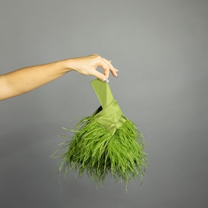 May include: A green satin clutch with a handle, adorned with vibrant green feathers. The bag is held against a gray background, showcasing its unique texture and color.