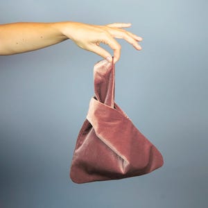 Może przedstawiać: Różowa aksamitna torebka z wiązanym uchwytem. Torebka ma trójkątny kształt i jest trzymana na jednolitym, szaro-niebieskim tle. Miękki, pluszowy materiał jest zebrany u góry, tworząc uchwyt.
