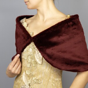 K&ouml;nnte beinhalten: Ein burgunderfarbener Kunstfellschal mit schlichtem Design. Der Schal ist &uuml;ber die Schultern einer Person gelegt, die ein goldenes Kleid tr&auml;gt.