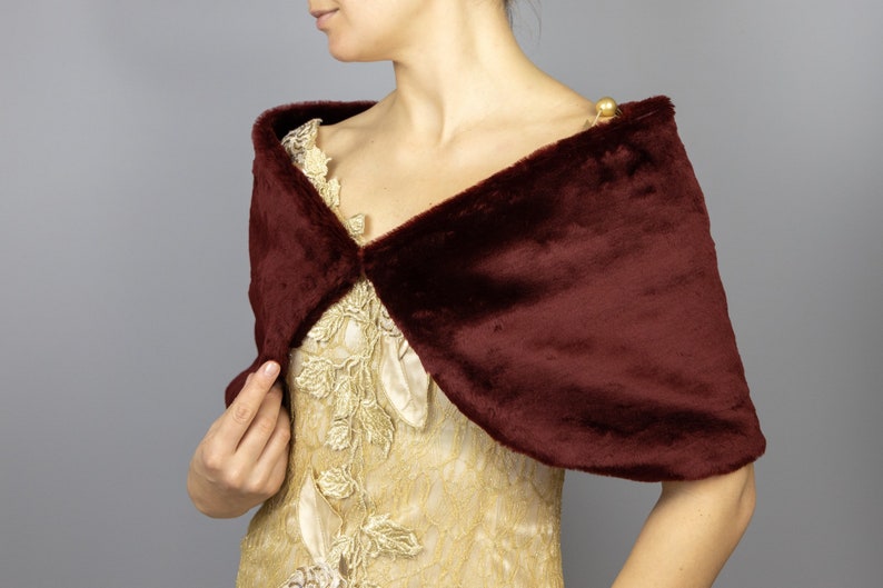 K&ouml;nnte beinhalten: Ein burgunderfarbener Kunstfellschal mit schlichtem Design. Der Schal ist &uuml;ber die Schultern einer Person gelegt, die ein goldenes Kleid tr&auml;gt.