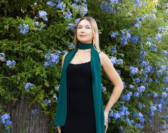 May include: A woman wearing a black dress and a teal scarf stands in front of a vibrant hedge of blue flowers. The scarf is draped around the neck and hangs down the front of the dress. The dress has a textured appearance.