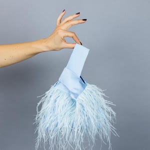 Peut inclure: Un sac à main bleu clair avec une poignée. Le sac est orné de douces plumes duveteuses de couleur bleu clair assortie. La poignée est en matière lisse, semblable à du satin. Le sac est posé sur un fond gris.