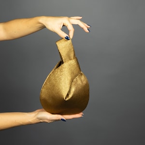 May include: A gold-coloured, knotted handbag with a soft, textured appearance. The bag is held up by two hands against a grey background. The bag's design features a unique knot detail at the top, creating a stylish and elegant look.