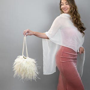 Puede incluir: Un bolso de mano color crema y con plumas, con un asa de cuentas, es sostenido. La persona lleva un chal blanco y una falda larga de terciopelo rosa empolvado. El fondo es gris liso.